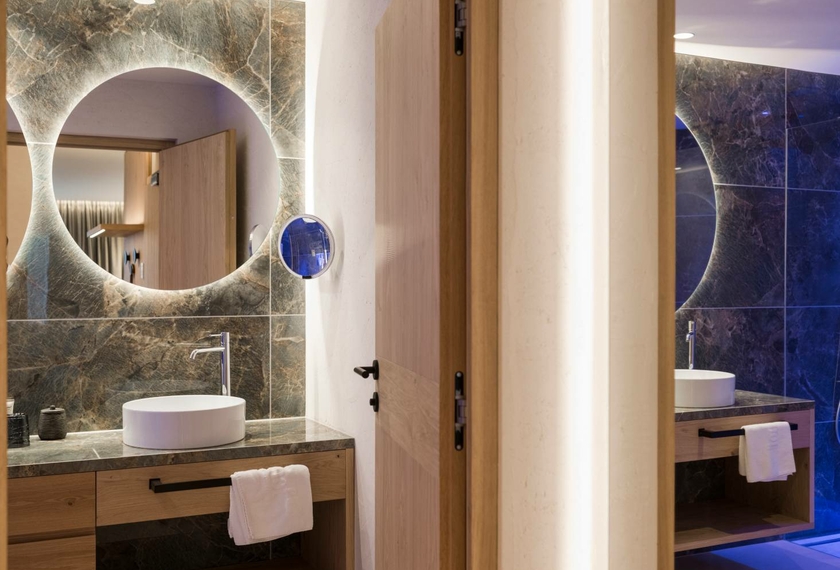 Doppelzimmer La Vita mit Bad im Feldhof Ein modernes Badezimmer mit rundem Spiegel, Waschtisch und Holztür neben dem Doppelzimmer La Vita.