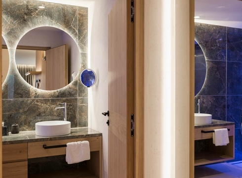 Doppelzimmer La Vita mit Bad im Feldhof Ein modernes Badezimmer mit rundem Spiegel, Waschtisch und Holztür neben dem Doppelzimmer La Vita.