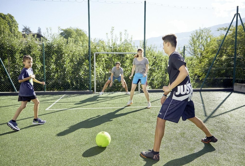 Familienurlaub Südtirol mit Fußballplatz Vier Kinder spielen auf einem kleinen Fußballplatz im Freien mit Bergblick.