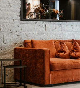 Ein orangefarbenes Loungesofa mit Kissen steht vor einer hellen Steinwand.
