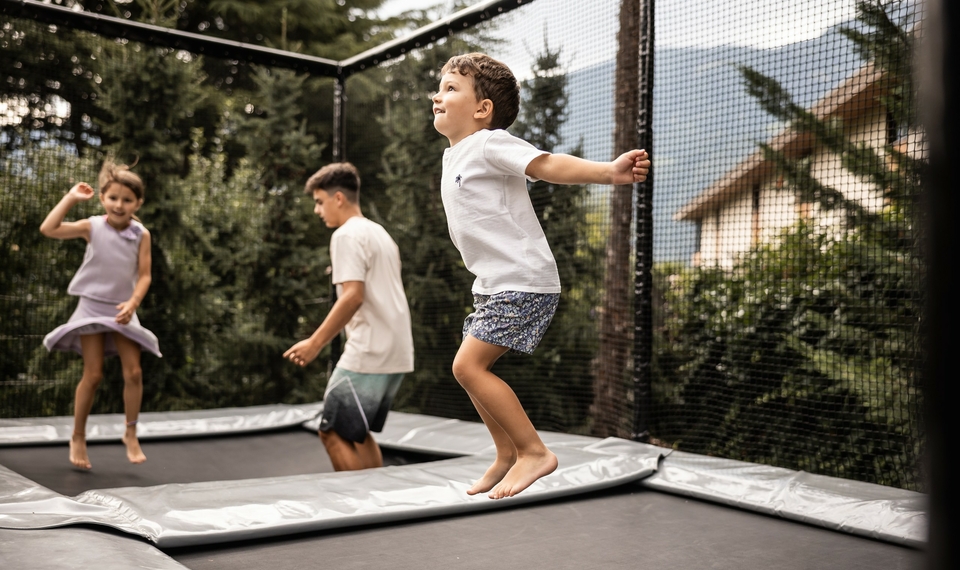 Familienurlaub in Südtirol auf dem Trampolin Drei Kinder springen gemeinsam auf einem Trampolin im Garten vor dem Hotel.