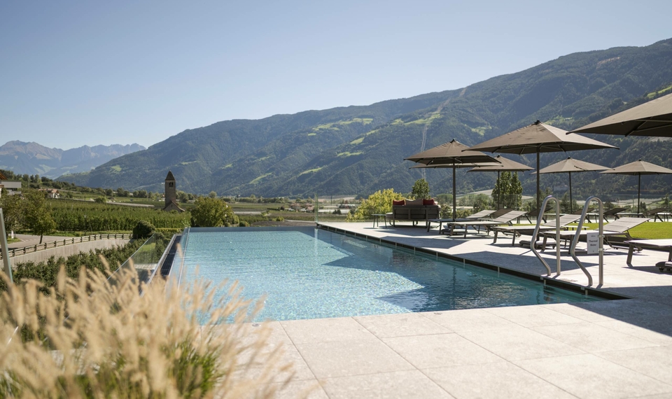 Ein Infinity-Pool mit Liegen und Sonnenschirmen liegt vor einer Bergkulisse.