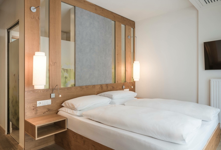 Das Doppelzimmer Spatzennest zeigt ein helles Bett mit Holzkopfteil, Spiegelwand und schlichter Einrichtung.