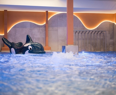 Innenpool mit Orca im Feldhof DolceVita Resort Ein Innenpool mit spritzendem Wasser und einer Orca-Figur im Feldhof DolceVita Resort.