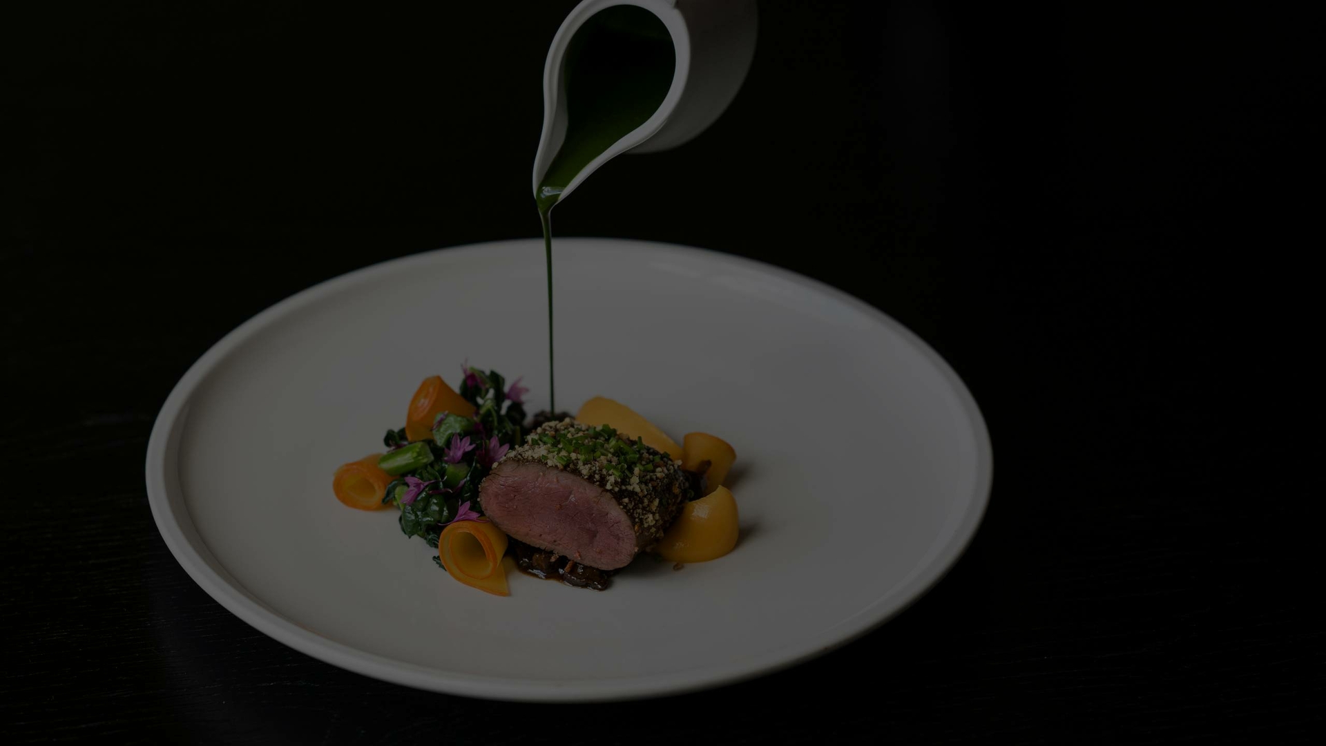 A plated gourmet dish on a white plate is being finished with sauce from a small jug.