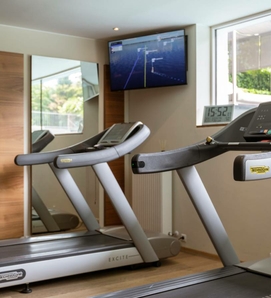 Zwei Laufbänder stehen vor einem Spiegel im Fitnessraum, darüber hängt ein Bildschirm an der Wand.