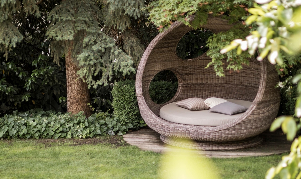 Ein runder Outdoor-Liegesessel mit Kissen steht im grünen Garten zwischen Bäumen und Sträuchern.
