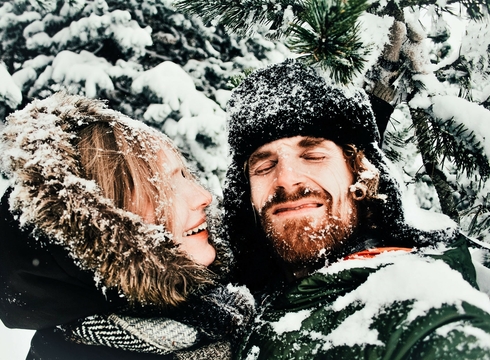 Winterurlaub im Schnee mit Paar im Wald Ein lächelndes Paar steht eng beieinander im verschneiten Wald und trägt Winterkleidung mit Fellkapuze.