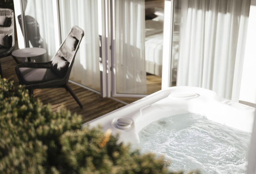 Whirlpool auf dem Balkon der Suite Bergzauber Ein Whirlpool auf dem Balkon mit Lounge-Sesseln und Blick durch helle Vorhänge ins Zimmer.