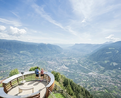 Zwei Personen stehen auf einer Aussichtsplattform mit weitem Blick über das Tal und die Berge.