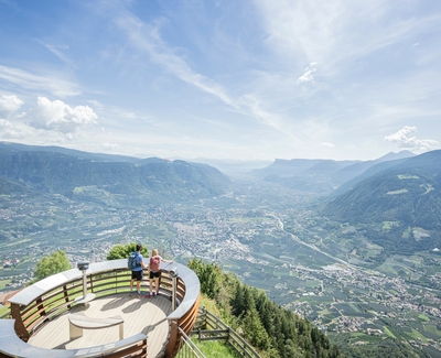 Due persone si trovano su una piattaforma panoramica con vista ampia sulla valle e sulle montagne.