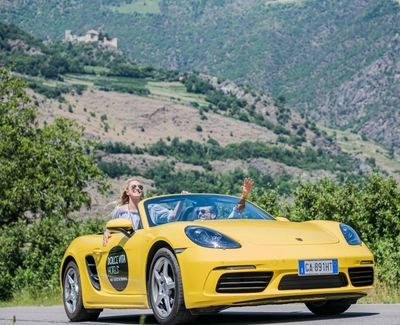 A yellow convertible drives on a road with two people and a green mountain backdrop.