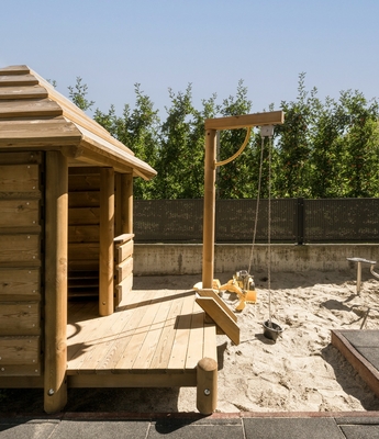 Kindersauna im Family Spa des Feldhof Eine kleine Holzhütte steht auf der Terrasse neben einem Sandspielbereich mit Spielgerät und Liegen.