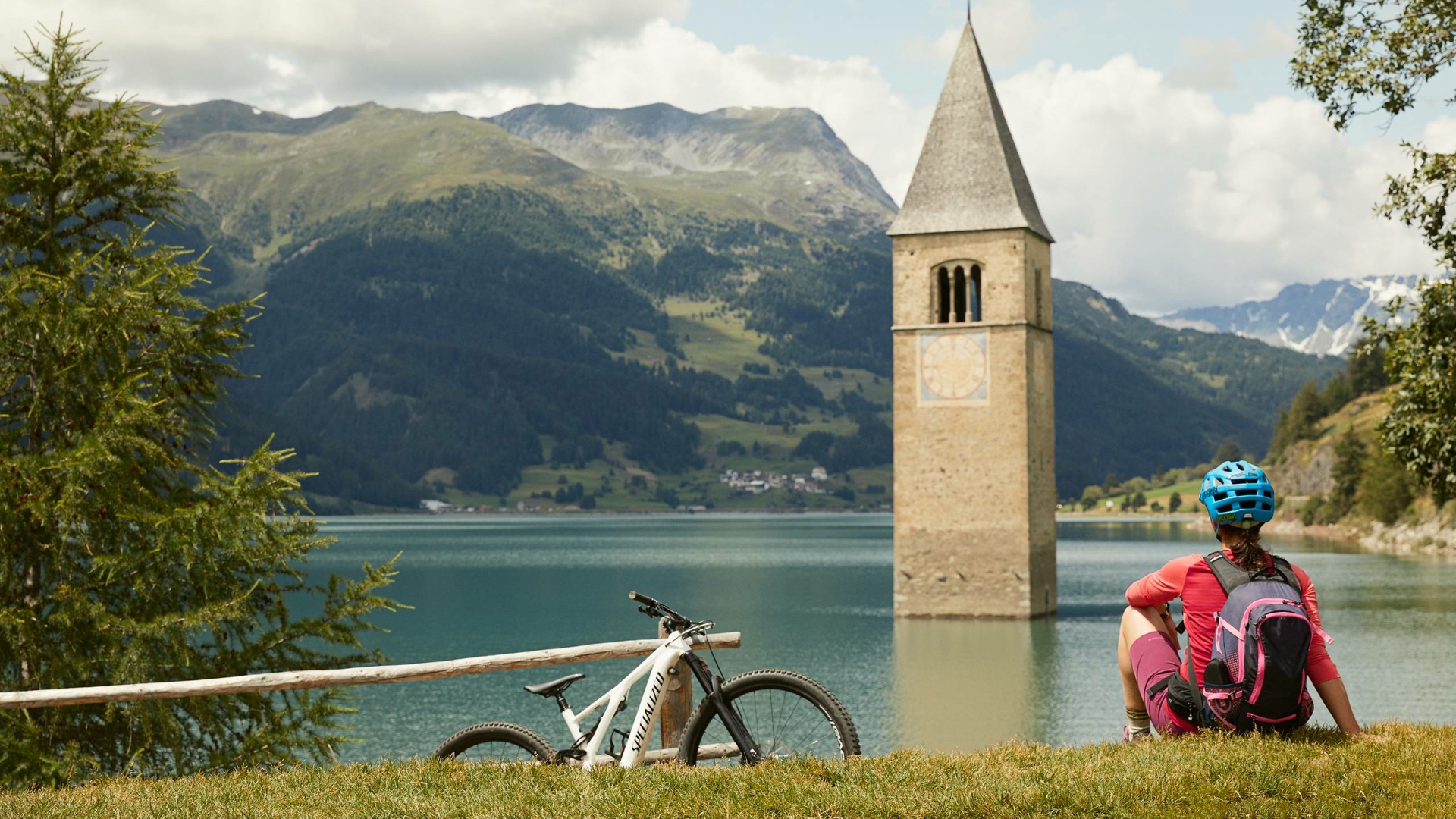A cyclist sits with a mountain bike on the lakeshore, facing a tower in the water and mountains behind it.