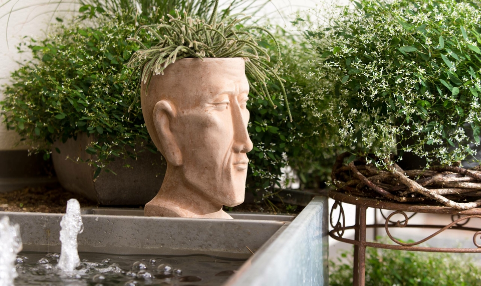 Ein Brunnen mit Wasserfontänen steht vor einer Pflanzenskulptur in einem Gartenbereich.