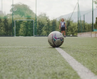 Fußballplatz im Hotel Feldhof in Naturns Ein Fußball liegt auf dem Kunstrasenplatz, im Hintergrund stehen zwei Kinder am Tor.