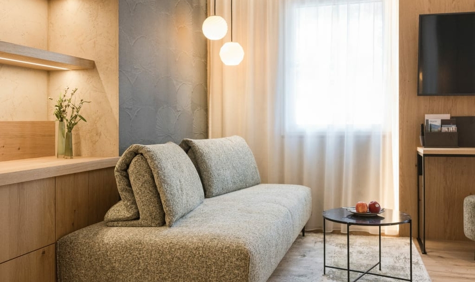Ein helles Hotelzimmer mit Sofa, kleinem Tisch und Fenster mit Vorhang.