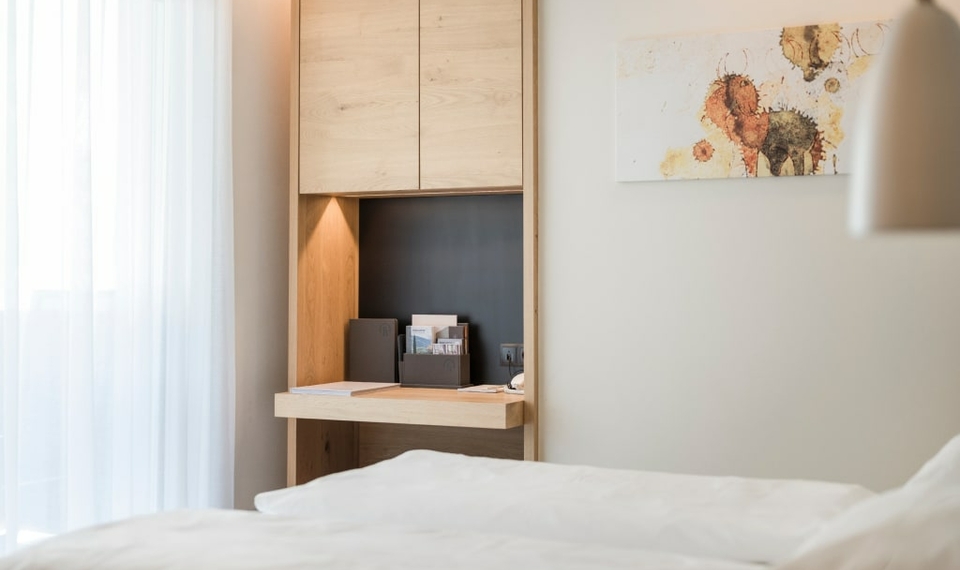 Ein helles Doppelzimmer mit Bett, Holzschreibtisch und Vorhang am Fenster.