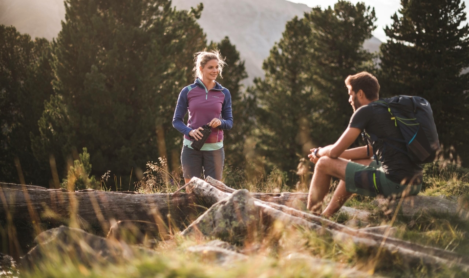 Eine Frau und ein Mann stehen beim Wandern auf einer Wiese mit Bäumen und Bergen im Hintergrund.