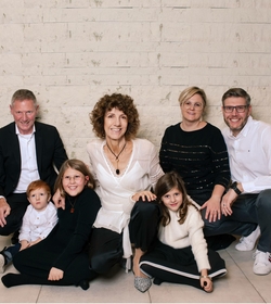 Sieben Personen einer Familie sitzen gemeinsam auf dem Boden vor einer hellen Steinwand.