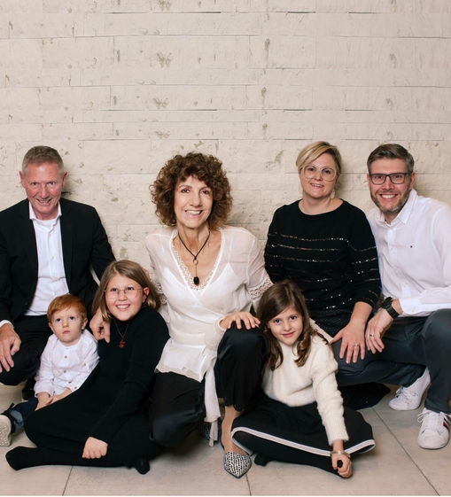 Sieben Personen einer Familie sitzen gemeinsam auf dem Boden vor einer hellen Steinwand.