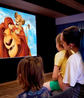 Kinderkino im Family Spa des Feldhof Drei Kinder sitzen vor einer Leinwand und sehen einen Löwenfilm im Kinderkino.
