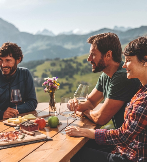 Drei Personen sitzen an einem Holztisch im Freien und genießen Essen und Wein mit Blick auf die Berge.