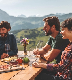 Drei Personen sitzen an einem Holztisch im Freien und genießen Essen und Wein mit Blick auf die Berge.