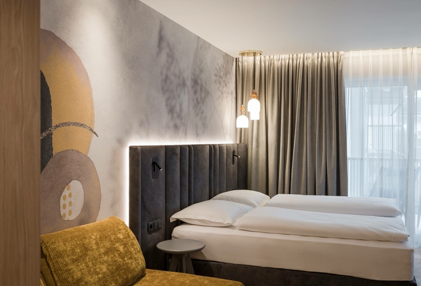 Doppelzimmer La Vita im Feldhof in Naturns Ein stilvoll eingerichtetes Doppelzimmer mit gepolstertem Bettkopfteil, Kunstwand und Vorhang am Fenster.