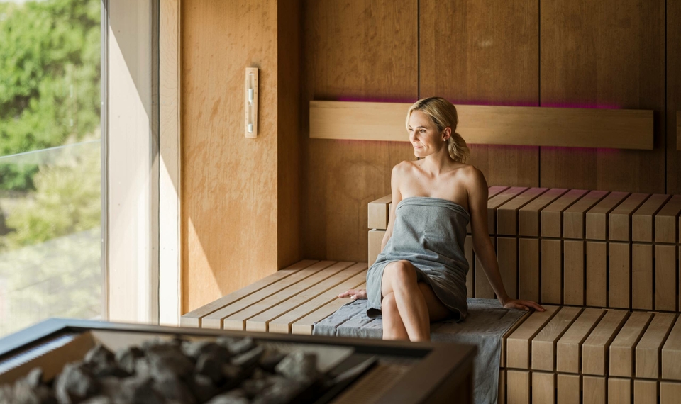 A woman sits on a wooden bench in a modern sauna and looks ahead calmly.