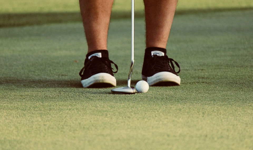 A golfer stands on the green with a club and ball in front of their shoes.
