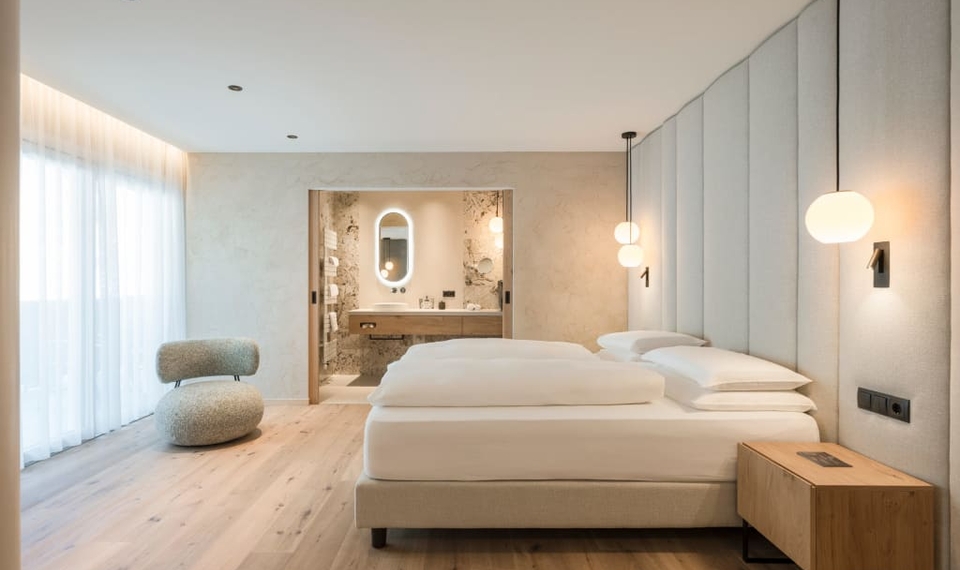 Ein helles Doppelzimmer mit großem Bett, gepolstertem Kopfteil, Holzdetails und Sitzbereich neben einem Badezimmer.