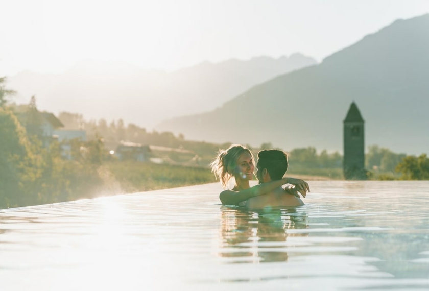 Infinity pool with mountain view at Feldhof A couple stands in the infinity pool, looking out toward the mountains in warm evening light.