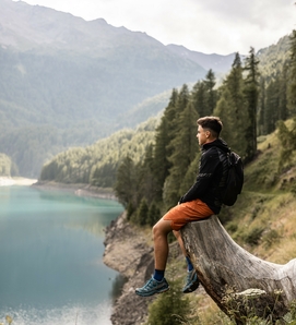 Ein Mann sitzt auf einem Baumstamm über einem Bergsee und blickt ins Tal.
