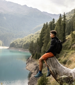 Ein Mann sitzt auf einem Baumstamm über einem Bergsee und blickt ins Tal.