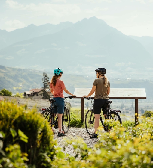 Two female cyclists stand with their e-bikes at a viewing table overlooking the mountains.