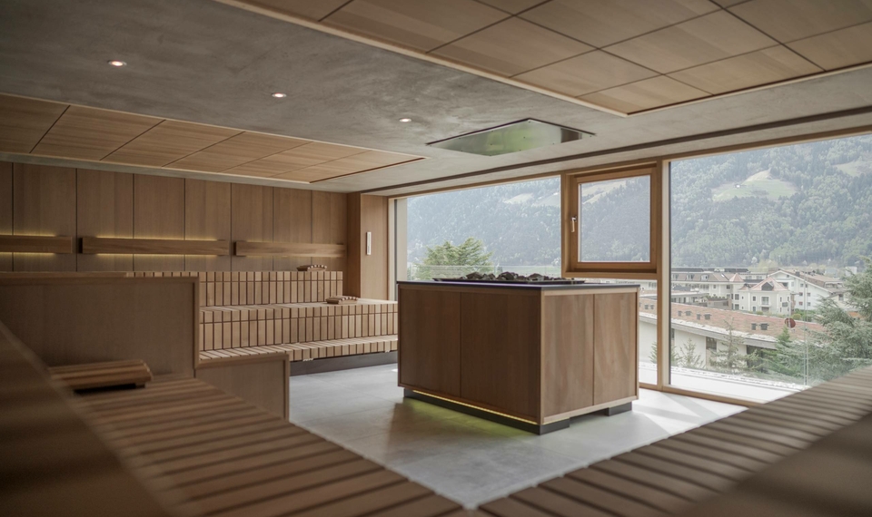 A bright sauna room with wooden loungers and views of the mountains and village through large windows.