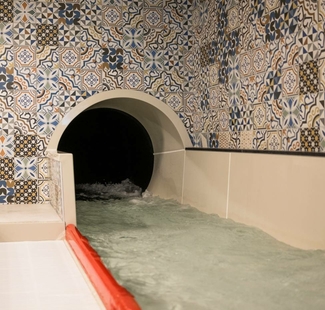 Eine Wasserrutsche führt in ein dunkles Becken, die Wände sind mit blau-beigen Fliesen verziert.