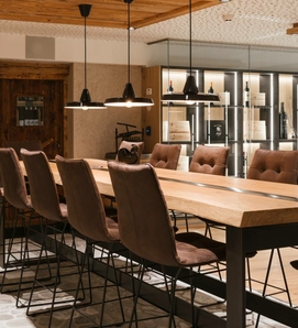 Ein langer Holztisch mit Barhockern steht vor einer beleuchteten Weintheke im Hotelrestaurant.