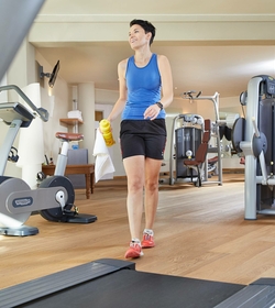 Eine Frau geht mit Handtuch und Trinkflasche durch den Fitnessraum.