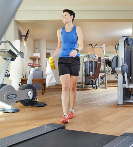 Eine Frau geht mit Handtuch und Trinkflasche durch den Fitnessraum.