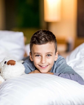 Ein Junge liegt lächelnd auf einem Bett und hält ein Kuscheltier im Familienzimmer.