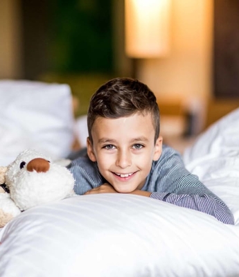 Camera familiare in hotel a Naturno Un ragazzo sorride sdraiato su un letto e tiene in mano un peluche nella camera familiare.