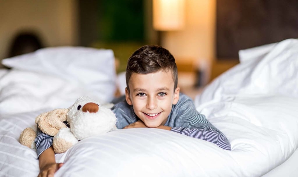 Familienzimmer im Hotel in Naturns Ein Junge liegt lächelnd auf einem Bett und hält ein Kuscheltier im Familienzimmer.