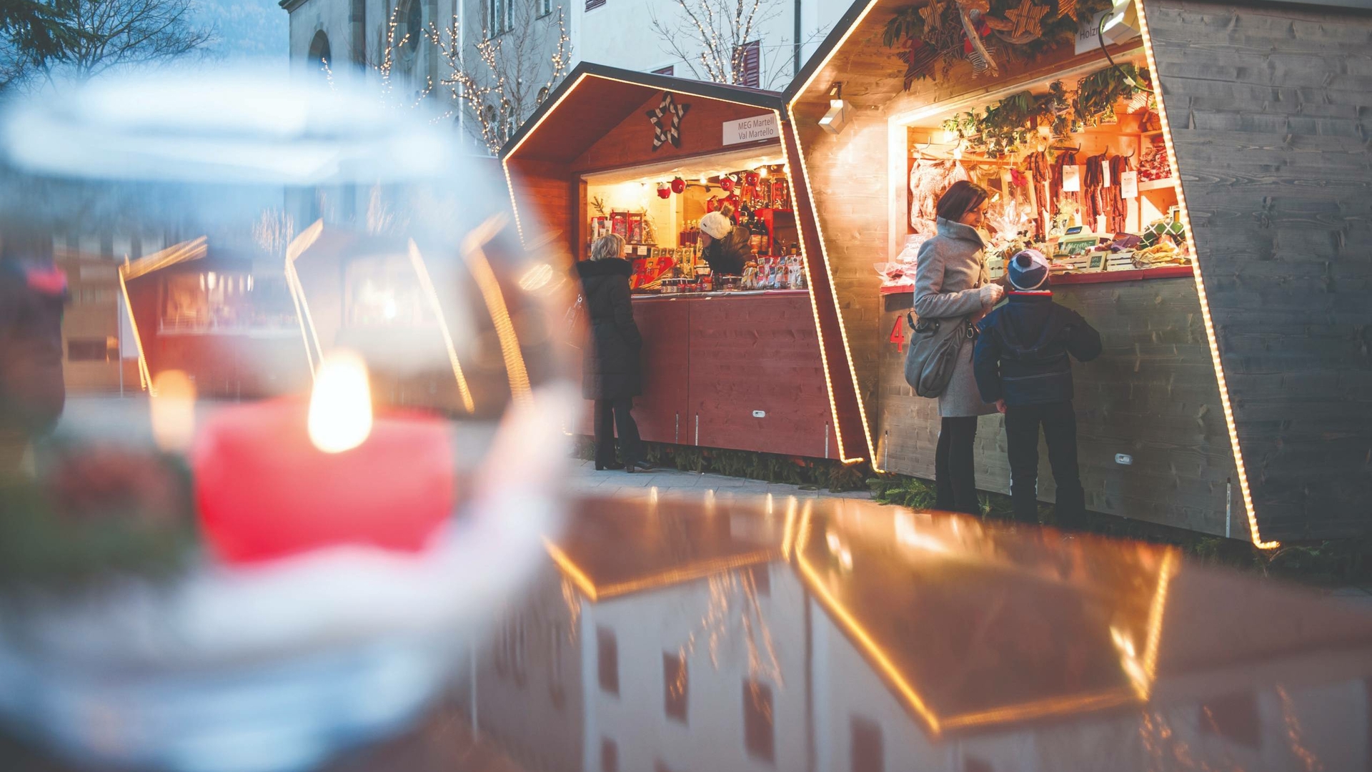 Due persone sono davanti a chalet di legno illuminati al mercatino di Natale, con una lanterna sfocata in primo piano.