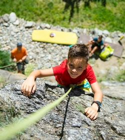 Ein Kind klettert gesichert an einem Felsen im Klettersteig, während weitere Personen im Hintergrund warten.