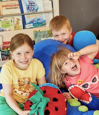 Kinderbetreuung im Familienhotel in Naturns Drei Kinder sitzen und liegen mit bunten Kissen in einem Spielbereich vor einem Regal mit Kinderbüchern.