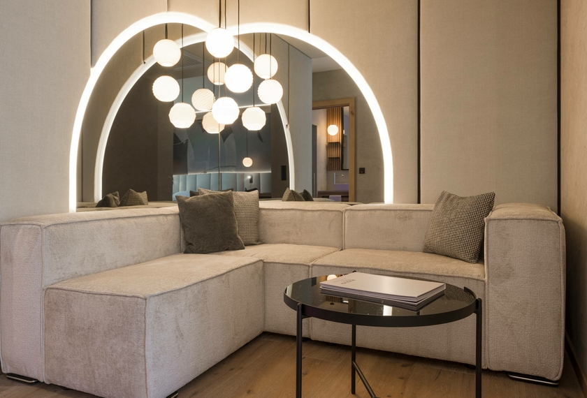 Luminoso soggiorno di una suite con divano angolare, tavolino rotondo e nicchia illuminata sullo sfondo.