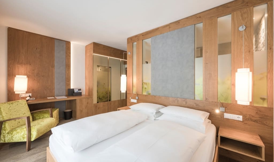 Ein Doppelbett steht in einem hellen Doppelzimmer mit Holzverkleidung und gepolstertem Sessel.