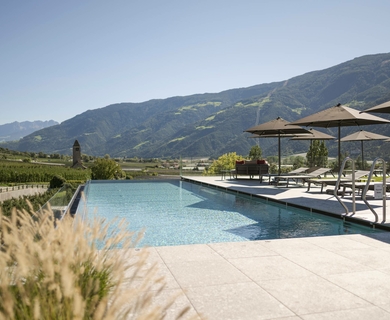 Poolterrasse im Feldhof DolceVita Resort in Naturns Der Außenpool mit Liegen und Sonnenschirmen liegt vor einer Bergkulisse.